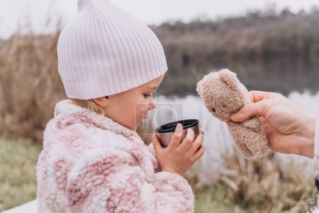 Téléchargez les photos : Bébé fille boire du thé d'un thermos près de la rivière à la fin de l'automne - en image libre de droit