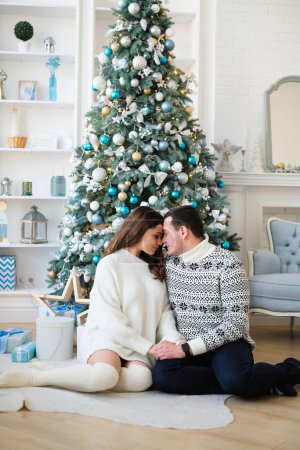 Photo for Couple is hugging near a Christmas tree with gifts in a room - Royalty Free Image