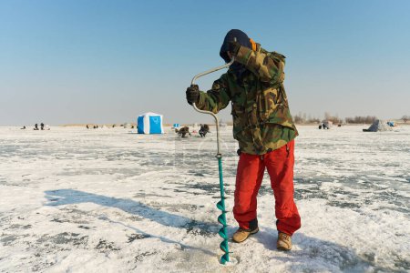 Photo for A warmly dressed Asian guy drills a hole in the ice on winter fi - Royalty Free Image