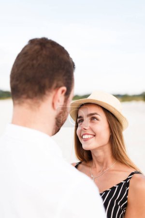 Téléchargez les photos : Young blonde woman in a hat looks at her partner on a sunny beach - en image libre de droit