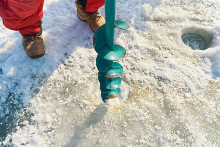 Foto de Un chico asiático calurosamente vestido taladra un agujero en el hielo en la fi de invierno - Imagen libre de derechos