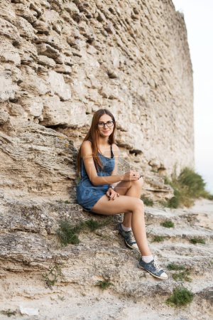 Foto de Mujer en overoles de mezclilla se sienta cerca de la pared de piedra en la montaña cerca de la ciudad - Imagen libre de derechos