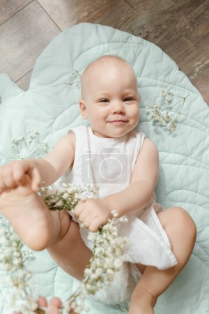 Photo for Bald baby girl in a white linen dress with gypsophila on a leaf rug - Royalty Free Image