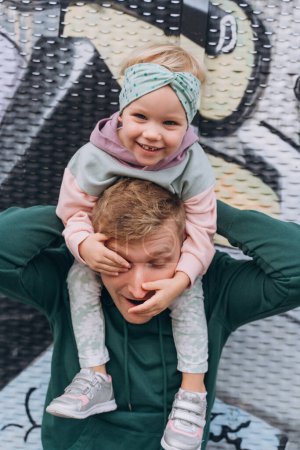 Téléchargez les photos : Élégant bébé fille est assis sur le cou de papa et rit, couleurs pastel - en image libre de droit