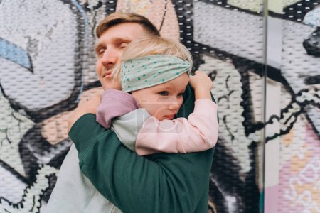 Téléchargez les photos : Papa en sweat à capuche étreignant sa petite fille devant des graffitis pastel - en image libre de droit