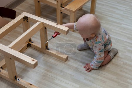 Foto de Niño pequeño sostiene destornillador cerca de los detalles de la silla de madera en el suelo - Imagen libre de derechos