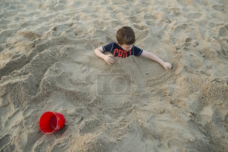 Téléchargez les photos : Vue d'en haut du garçon enterré jusqu'à sa poitrine dans le sable à la plage - en image libre de droit