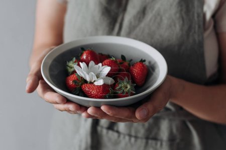 Foto de Un plato de cerámica gris con fresas y flor blanca en las manos. - Imagen libre de derechos