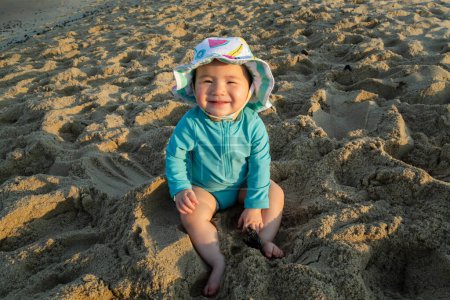 Téléchargez les photos : Bébé fille souriante heureuse portant un maillot de bain sourit tout en étant assis sur le sable - en image libre de droit