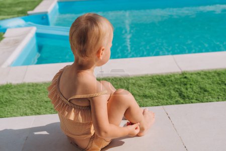 Foto de Bebé en traje de baño se sienta en un césped sintético cerca de la piscina - Imagen libre de derechos