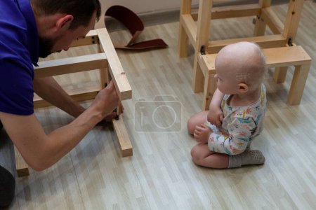 Foto de Niño se sienta de rodillas cerca de padre construyendo silla de madera para él - Imagen libre de derechos