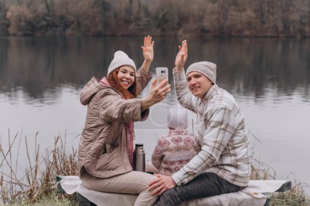 Téléchargez les photos : Une famille s'assoit au bord de la rivière dans le parc d'automne et fait un selfie - en image libre de droit