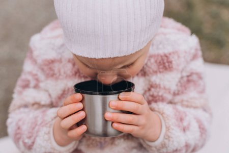 Téléchargez les photos : Bébé fille boire du thé d'un thermos près de la rivière à la fin de l'automne - en image libre de droit