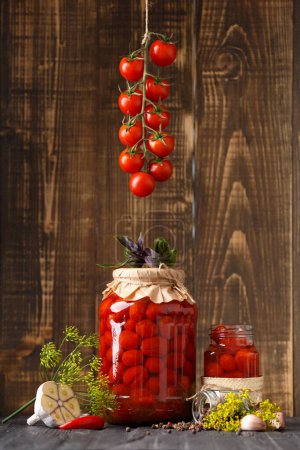 Photo for Pickled cherry tomatoes with garlic, dill and spices. Homemade food. Canned tomatoes in closed and open jars with a branch of fresh cherry tomatoes on a wooden background. Pickled vegetables. - Royalty Free Image