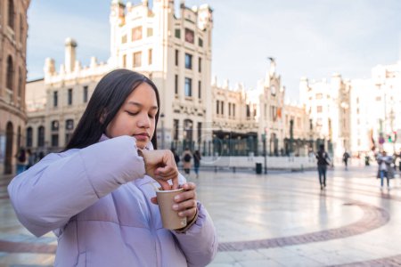 Foto de Chica con taza de chocolate de papel en el centro de la ciudad sumergiendo algo. - Imagen libre de derechos