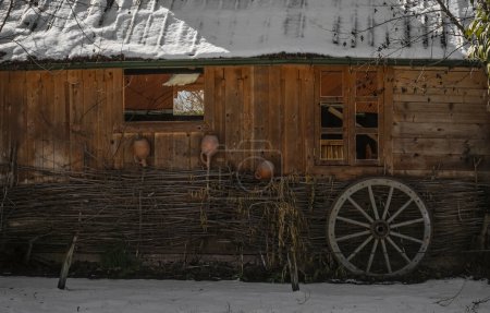 Téléchargez les photos : Façade de l'ancienne maison en bois rond. Maison de village géorgien vieille fenêtre avec ancienne roue de calèche. cruche à vin en céramique sur la clôture. journée d'hiver enneigée - en image libre de droit