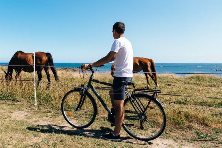 Téléchargez les photos : Attrayant jeune homme équitation vélo dans l'île de Batz avec des chevaux - en image libre de droit