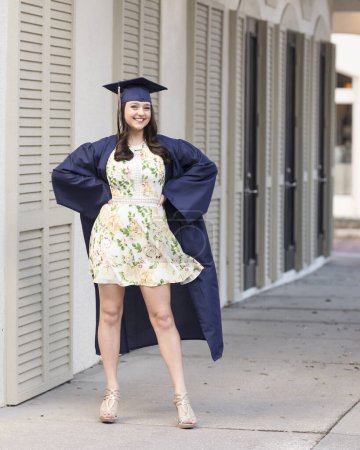 Photo for Graduating girl in cap and gown and floral dress poses on street - Royalty Free Image