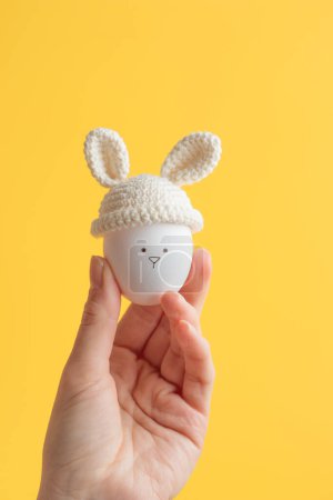 Téléchargez les photos : Œuf de Pâques au crochet chapeau avec des oreilles de lapin dans une main humaine sur fond jaune. Bannière verticale, carte postale avec espace de copie - en image libre de droit