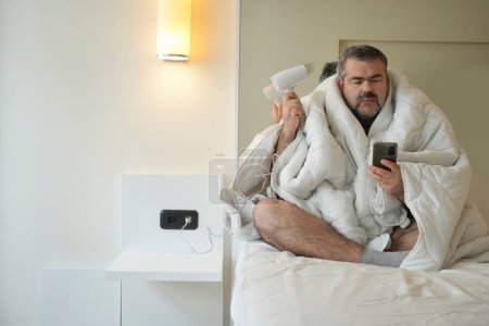 Téléchargez les photos : Moyen âge mâle assis sur le lit sous couverture blanche, l'homme en utilisant smartphone et sèche-cheveux - en image libre de droit
