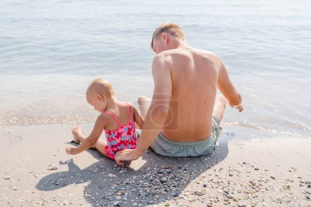 Foto de Niña bebé niño con papá sentarse en la playa junto al mar, vista trasera - Imagen libre de derechos