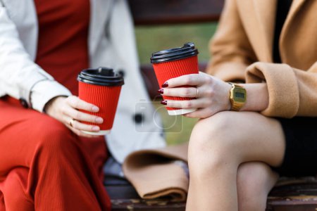 Photo for Two women holding paper cups with hot drinks sitting on a bench - Royalty Free Image