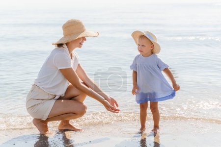 Téléchargez les photos : Maman avec une petite fille en chapeaux de paille sur la plage au bord de la mer - en image libre de droit