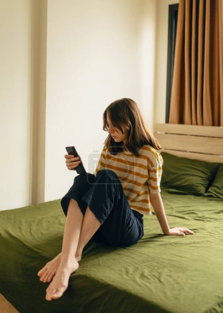 Foto de Mujer joven mirando su teléfono mientras está sentada en una cama - Imagen libre de derechos