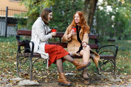 Téléchargez les photos : Filles s'amuser assis avec des boissons chaudes dans le parc d'automne sur un banc - en image libre de droit
