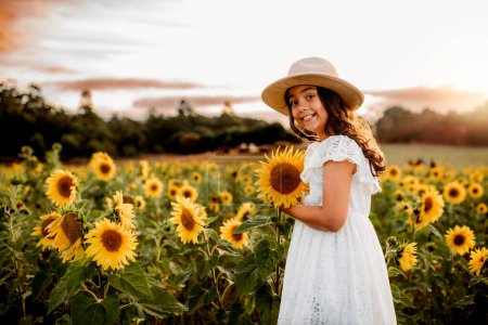 Photo for Young  girl with a hat on holding a sunflower in a field at sunset - Royalty Free Image