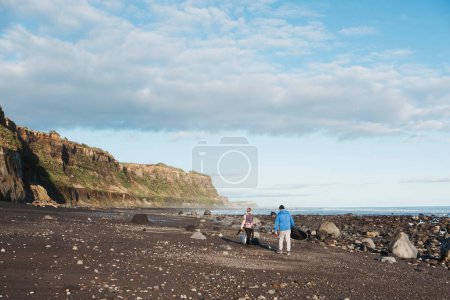 Téléchargez les photos : Père portant une planche de surf et fils promenant un chien sur une plage rocheuse - en image libre de droit