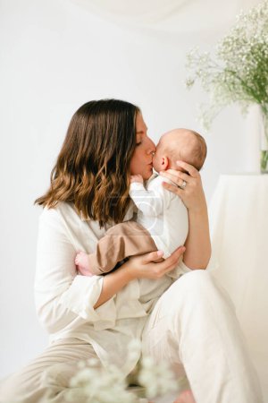 Sitting young mother kissing newborn baby son by baby's breath flowers