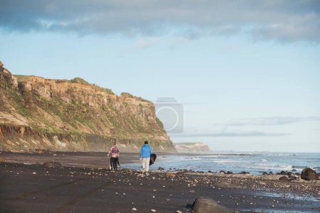 Photo for Father and son walking down rocky beach with a surfboard - Royalty Free Image