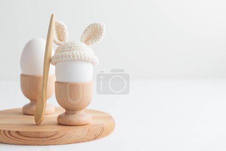 Foto de Huevo de Pascua en sombrero de ganchillo con orejas de conejo en soporte de huevo de madera sobre fondo blanco. Banner horizontal, tarjeta postal con copyspace - Imagen libre de derechos