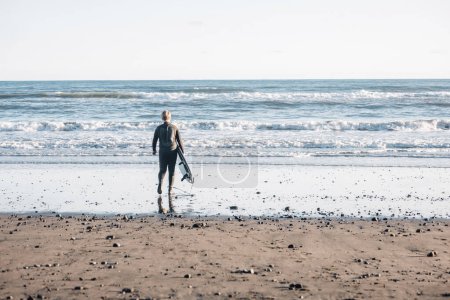 Photo for Tween boy in a wetsuit holding a surfboard walking into the water - Royalty Free Image