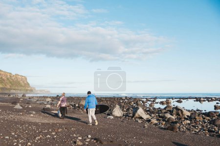 Téléchargez les photos : Père et fils promenant un chien sur une plage rocheuse portant une planche de surf - en image libre de droit