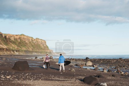 Téléchargez les photos : Père et fils marchant sur la plage rocheuse avec une planche de surf et un chien - en image libre de droit