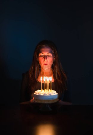 Photo for Attractive woman blowing out candles on birthday cake in dark room. - Royalty Free Image
