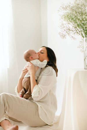 Téléchargez les photos : Assis jeune mère embrasser le nouveau-né fils par la fenêtre et les fleurs - en image libre de droit