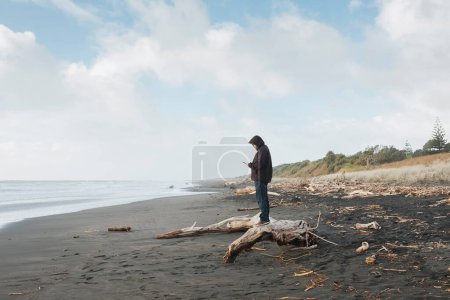 Téléchargez les photos : Homme debout sur le bois flotté regardant le téléphone portable à la plage - en image libre de droit