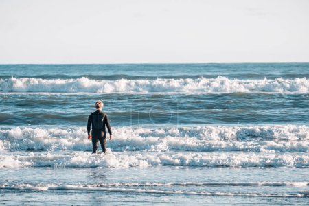 Téléchargez les photos : Entre garçon portant une combinaison debout dans le surf à la plage - en image libre de droit