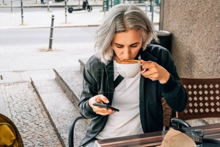 Téléchargez les photos : Femme boit du café et utilise le téléphone à la table du café extérieur - en image libre de droit