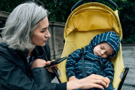 Téléchargez les photos : Femme regarde dormir enfant assis dans une poussette avec téléphone dans les mains - en image libre de droit