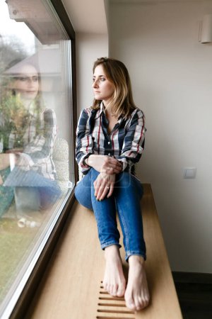 Foto de Mujer joven en ropa de casa se sienta sola en el alféizar de la ventana en pensamiento - Imagen libre de derechos