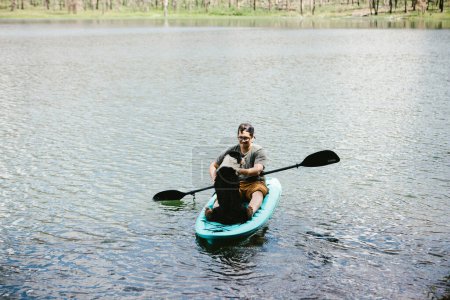 Téléchargez les photos : Homme et chien dans un kayak sarcelle sur un lac avec forêt derrière eux. - en image libre de droit
