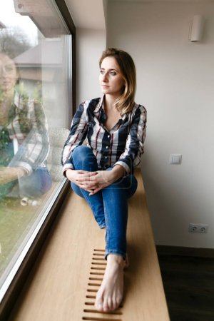 Téléchargez les photos : Jeune femme en vêtements de maison est assis seul sur le rebord de la fenêtre dans la pensée - en image libre de droit