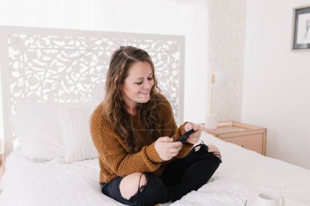 Téléchargez les photos : Heureuse jeune femme regardant le téléphone assis sur un lit blanc - en image libre de droit