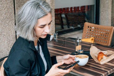 Téléchargez les photos : Femme aux cheveux gris est assis au café de la rue et utilise un smartphone - en image libre de droit