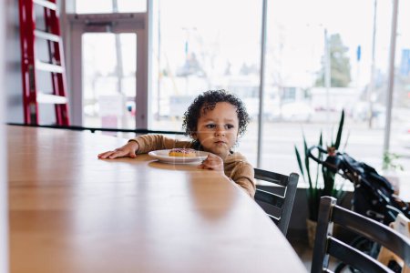 Foto de Niño pensativo sentado en la mesa de la cafetería con rosquilla - Imagen libre de derechos