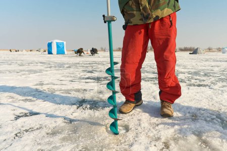 Photo for A warmly dressed Asian guy drills a hole in the ice on winter fishing, body part - Royalty Free Image